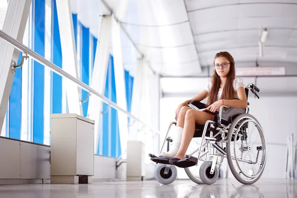 Uma rapariga numa cadeira de rodas está a ler um livro. Paciente em cadeira de rodas no corredor do hospital — Fotografia de Stock