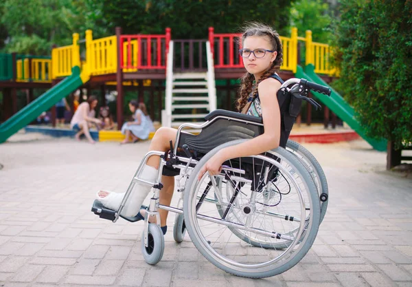 Девушка со сломанной ногой сидит в инвалидном кресле перед детской площадкой — стоковое фото