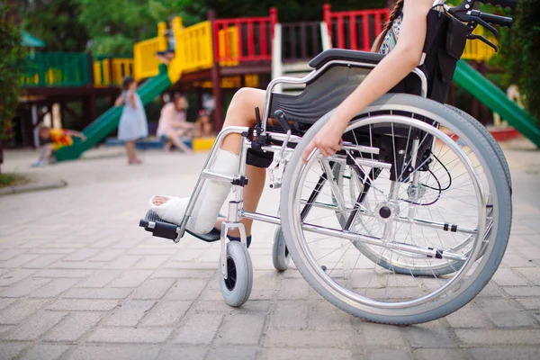 Uma menina com uma perna quebrada senta-se em uma cadeira de rodas na frente do playground — Fotografia de Stock