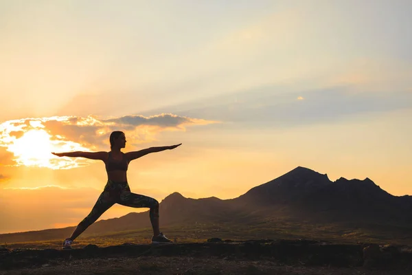 Silueta de mujer joven practicando yoga o pilates al atardecer o al amanecer en una hermosa ubicación de montaña, haciendo ejercicio de embestida, de pie en Warrior — Foto de Stock