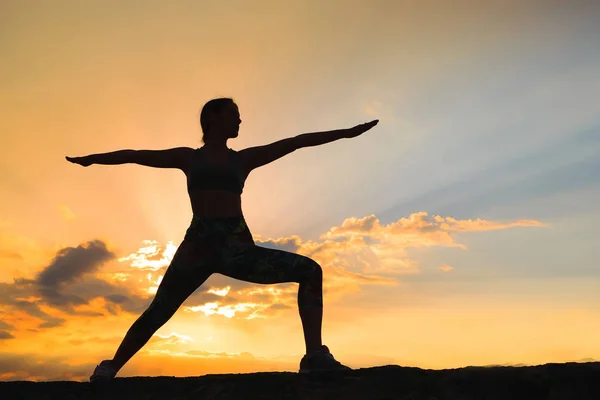 Silueta de mujer joven practicando yoga o pilates al atardecer o al amanecer en una hermosa ubicación de montaña, haciendo ejercicio de embestida, de pie en Warrior — Foto de Stock