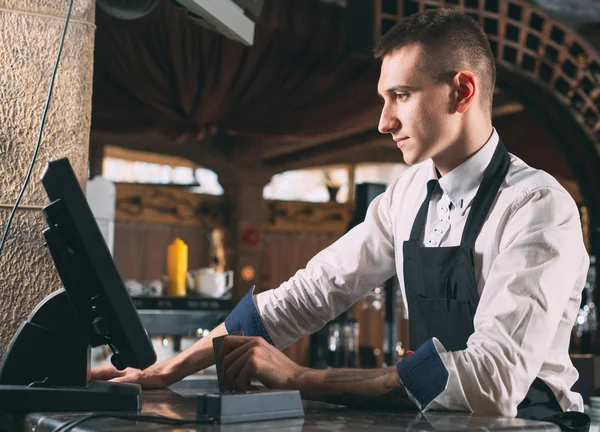 small business, people and service concept - happy man or waiter in apron at counter with cashbox working at bar or coffee shop.