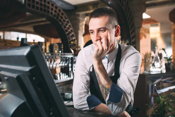 small business, people and service concept - happy man or waiter in apron at counter with cashbox working at bar or coffee shop.