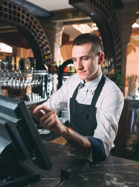 small business, people and service concept - happy man or waiter in apron at counter with cashbox working at bar or coffee shop.