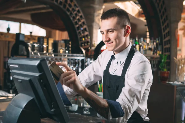 small business, people and service concept - happy man or waiter in apron at counter with cashbox working at bar or coffee shop.