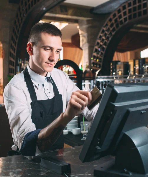 small business, people and service concept - happy man or waiter in apron at counter with cashbox working at bar or coffee shop