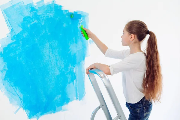 Repair in the apartment. Happy child girl paints the wall with blue paint, — Stock Photo, Image