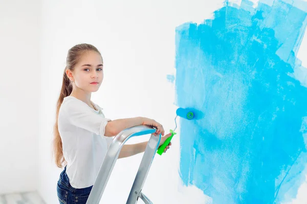 Repair in the apartment. Happy child girl paints the wall with blue paint, — Stock Photo, Image