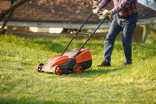 Maaimachine - werknemer maait gras in groene tuin bij zonsondergang. Man met elektrische grasmaaier, grasmaaier. Tuinier trimmen een tuin. — Stockfoto