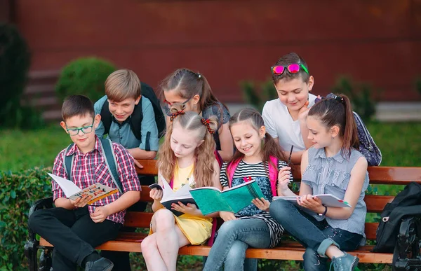 Счастливый школьный портрет. Одноклассники сидят с книгами на деревянной скамейке в городском парке и учатся в солнечный день. — стоковое фото