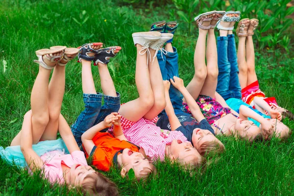 Παιδιά ξαπλωμένα στο πράσινο γρασίδι στο πάρκο μια καλοκαιρινή μέρα με τα πόδια τους σηκωμένα στον ουρανό. — Φωτογραφία Αρχείου
