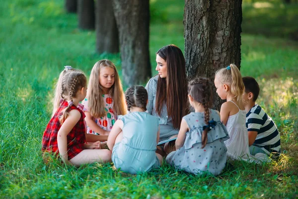 Children and education, young woman at work as educator reading book to boys and girls in park. — Stock Photo, Image
