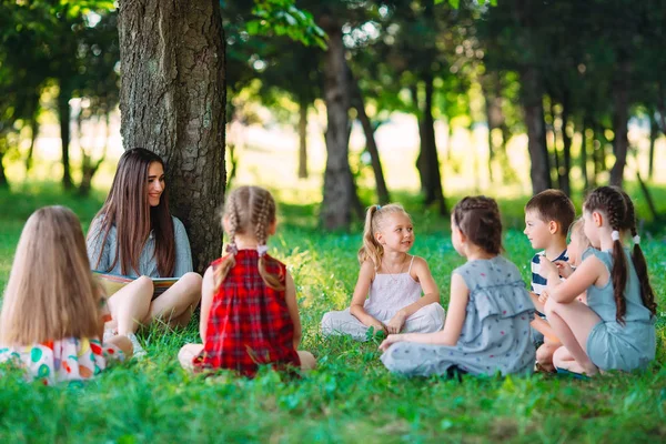 Children and education, young woman at work as educator reading book to boys and girls in park. — Stock Photo, Image