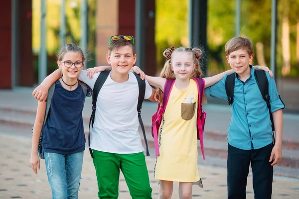 Amistad infantil. Cuatro pequeños estudiantes de la escuela, dos niños y dos niñas, de pie en un abrazo en el patio de la escuela. — Foto de Stock