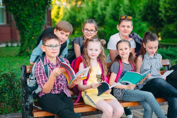 Счастливый школьный портрет. Одноклассники сидят с книгами на деревянной скамейке в городском парке и учатся в солнечный день. — стоковое фото