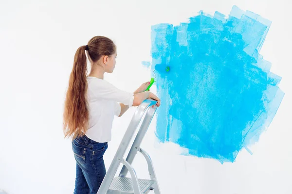 Repair in the apartment. Happy child girl paints the wall with blue paint, — Stock Photo, Image