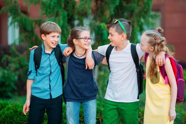 Amistad infantil. Cuatro pequeños estudiantes de la escuela, dos niños y dos niñas, de pie en un abrazo en el patio de la escuela. — Foto de Stock