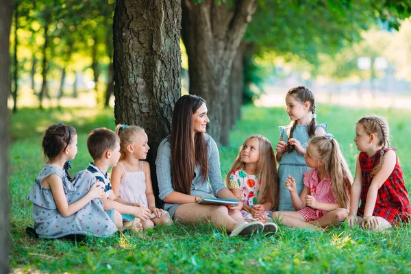 Children and education, young woman at work as educator reading book to boys and girls in park. — Stock Photo, Image