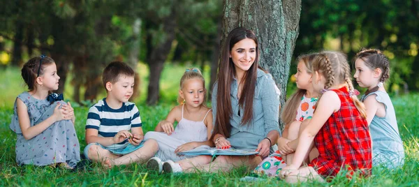 Niños y educación, mujer joven en el trabajo como educadora leyendo libro para niños y niñas en el parque. — Foto de Stock