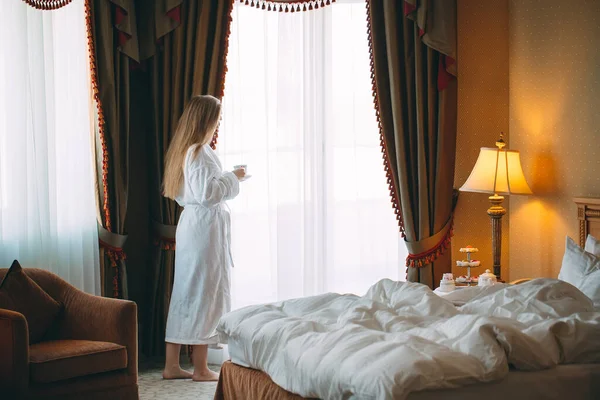 La chica Stit cerca de la ventana y bebe café de la mañana en la habitación del hotel o en casa. —  Fotos de Stock