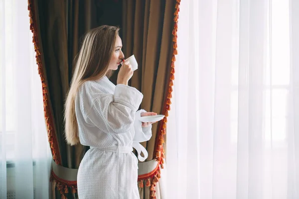 Το κορίτσι Stit κοντά στο παράθυρο και πίνει πρωινό καφέ στο δωμάτιο του ξενοδοχείου ή στο σπίτι. — Φωτογραφία Αρχείου
