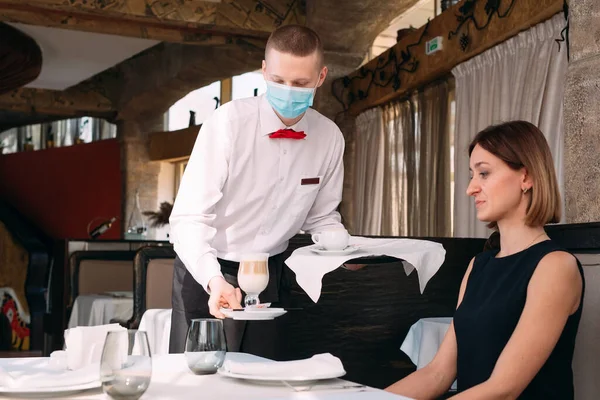 A European-looking waiter in a medical mask serves Latte coffee. — Stock Photo, Image