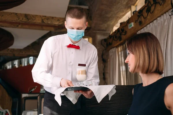 A European-looking waiter in a medical mask serves Latte coffee. — Stock Photo, Image