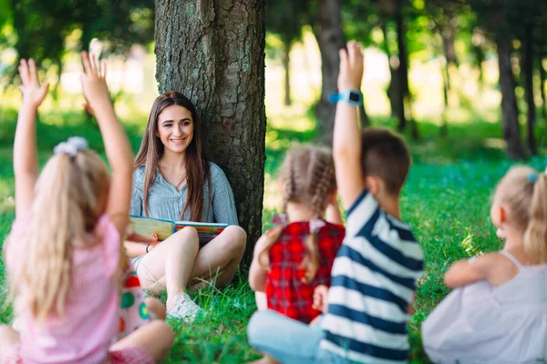 Niños y educación, mujer joven en el trabajo como educadora leyendo libro para niños y niñas en el parque. —  Fotos de Stock