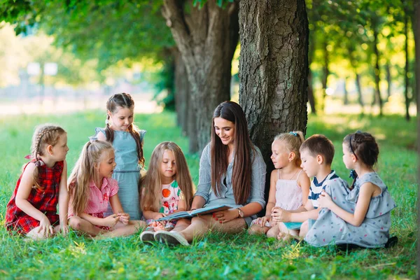 Bambini e istruzione, giovane donna al lavoro come educatrice lettura libro a ragazzi e ragazze nel parco. — Foto Stock