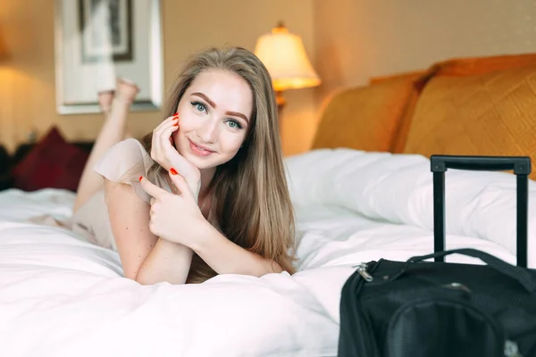 Smiling wanita bisnis muda di tempat tidur menatap kamera. Kamar hotel. Gadis itu menjilat di tempat tidur dekat koper. — Stok Foto