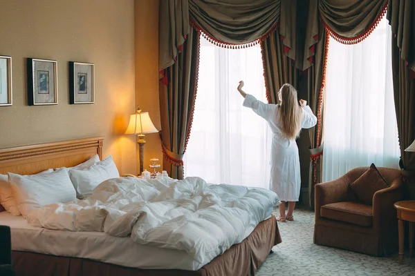 Mujer en albornoz estancia cerca de la ventana en la habitación del hotel. —  Fotos de Stock