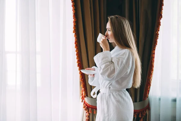 La chica Stit cerca de la ventana y bebe café de la mañana en la habitación del hotel o en casa. —  Fotos de Stock