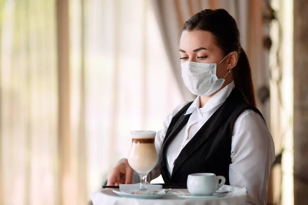 A female Waiter of European appearance in a medical mask serves Latte coffee. — Stock Photo, Image