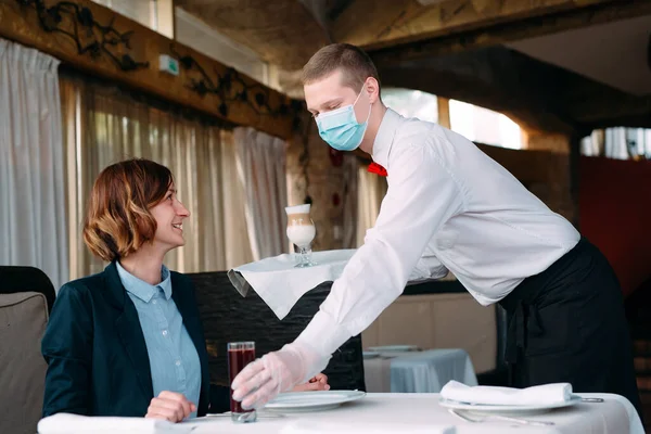 A European-looking waiter in a medical mask serves Latte coffee. — Stock Photo, Image