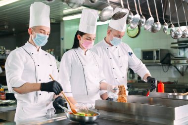 Koruyucu maskeli ve eldivenli aşçılar bir restoranın ya da otelin mutfağında yemek hazırlıyorlar..