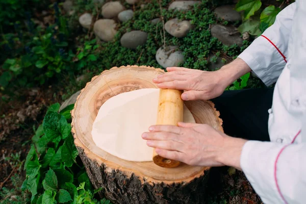 A restaurant chef in uniform rolls out dough on a stump in the woods.