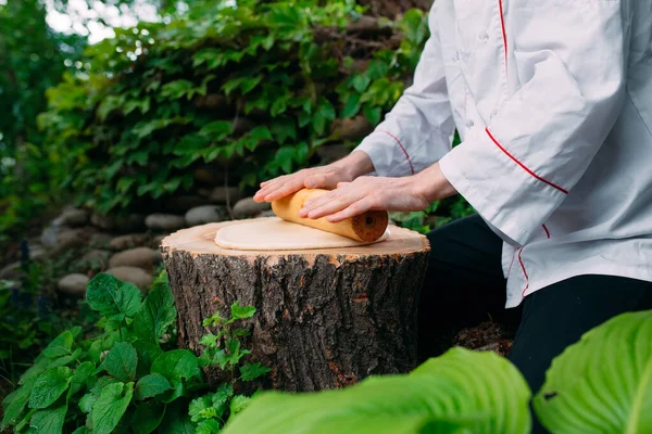 A restaurant chef in uniform rolls out dough on a stump in the woods.
