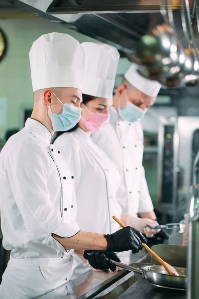 Koks in beschermende maskers en handschoenen bereiden voedsel in de keuken van een restaurant of hotel. — Stockfoto