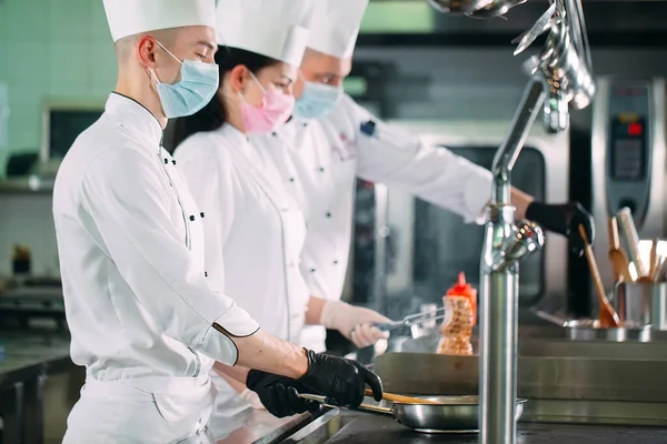 Chefs con máscaras protectoras y guantes preparan comida en la cocina de un restaurante u hotel. — Foto de Stock