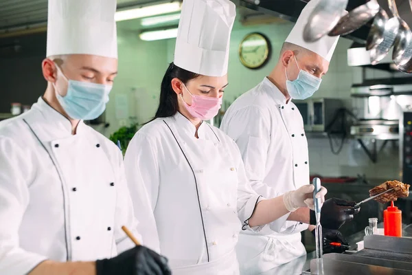 Chefs con máscaras protectoras y guantes preparan comida en la cocina de un restaurante u hotel. — Foto de Stock