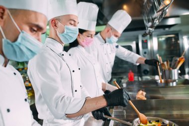 Koruyucu maskeli ve eldivenli aşçılar bir restoranın ya da otelin mutfağında yemek hazırlıyorlar..