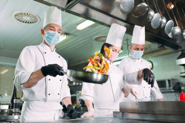 Koruyucu maskeli ve eldivenli aşçılar bir restoranın ya da otelin mutfağında yemek hazırlıyorlar..