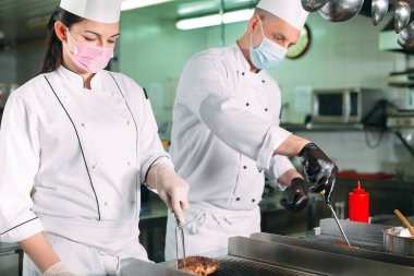 Koruyucu maskeli ve eldivenli aşçılar bir restoranın ya da otelin mutfağında yemek hazırlıyorlar..