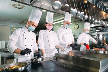Koruyucu maskeli ve eldivenli aşçılar bir restoranın ya da otelin mutfağında yemek hazırlıyorlar..