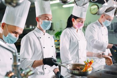Koruyucu maskeli ve eldivenli aşçılar bir restoranın ya da otelin mutfağında yemek hazırlıyorlar..