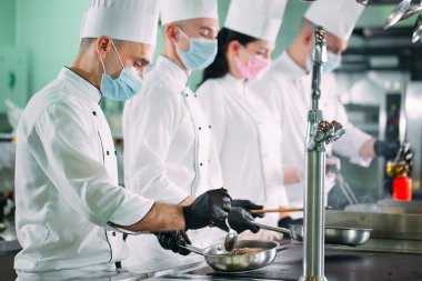 Koruyucu maskeli ve eldivenli aşçılar bir restoranın ya da otelin mutfağında yemek hazırlıyorlar..