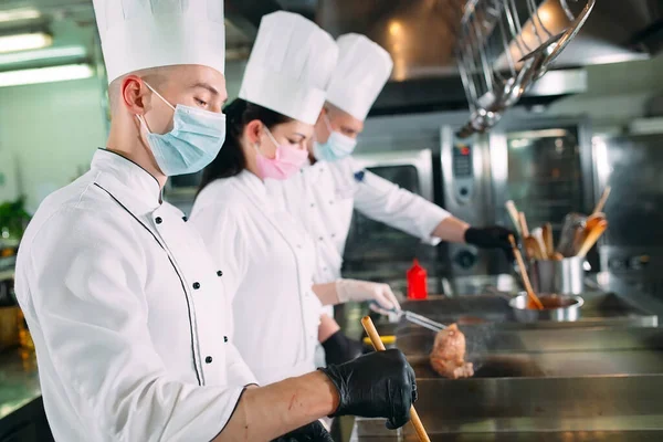 Les chefs masqués et les gants de protection préparent la nourriture dans la cuisine d'un restaurant ou d'un hôtel. — Photo