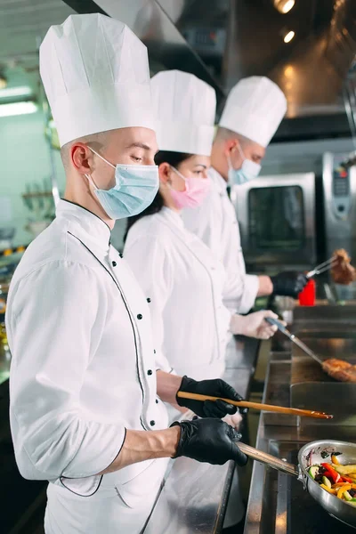 Koks in beschermende maskers en handschoenen bereiden voedsel in de keuken van een restaurant of hotel. — Stockfoto