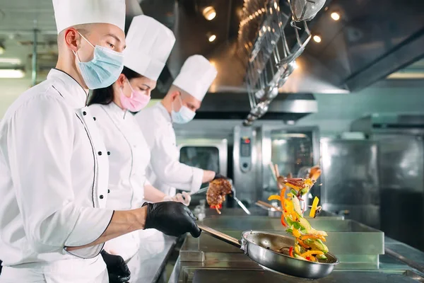 Chefs con máscaras protectoras y guantes preparan comida en la cocina de un restaurante u hotel. —  Fotos de Stock