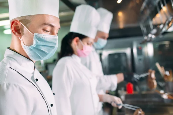 Chefs con máscaras protectoras y guantes preparan comida en la cocina de un restaurante u hotel. —  Fotos de Stock